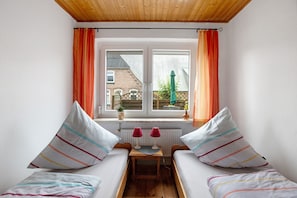 Una habitación pequeña con dos camas individuales, una mesa con dos lámparas, una ventana con cortinas y vistas a un edificio de ladrillo.