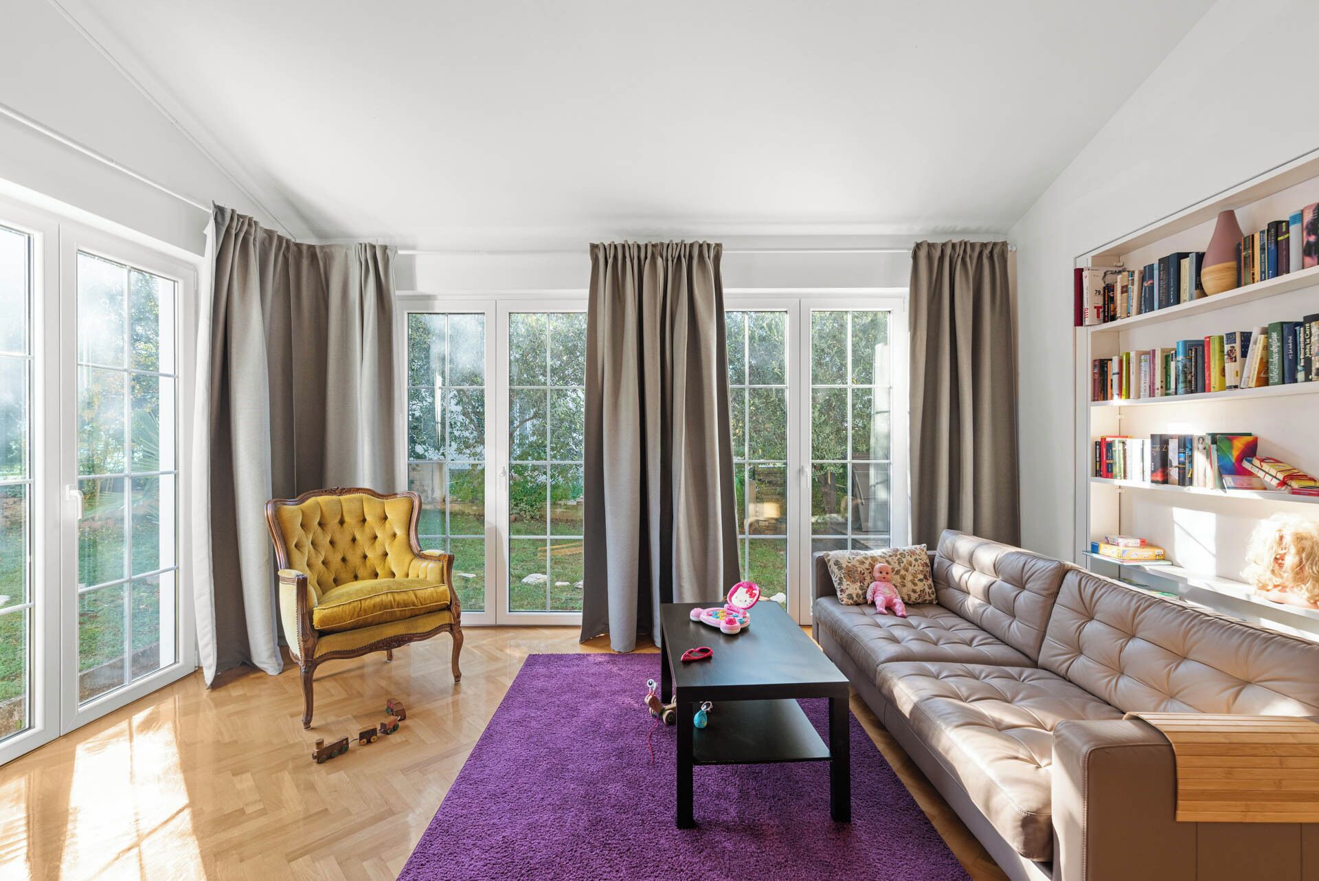 Ein geräumiges Wohnzimmer mit großen Fenstern, einem gelben Sessel, einer Couch, einem Couchtisch und einem Bücherregal.