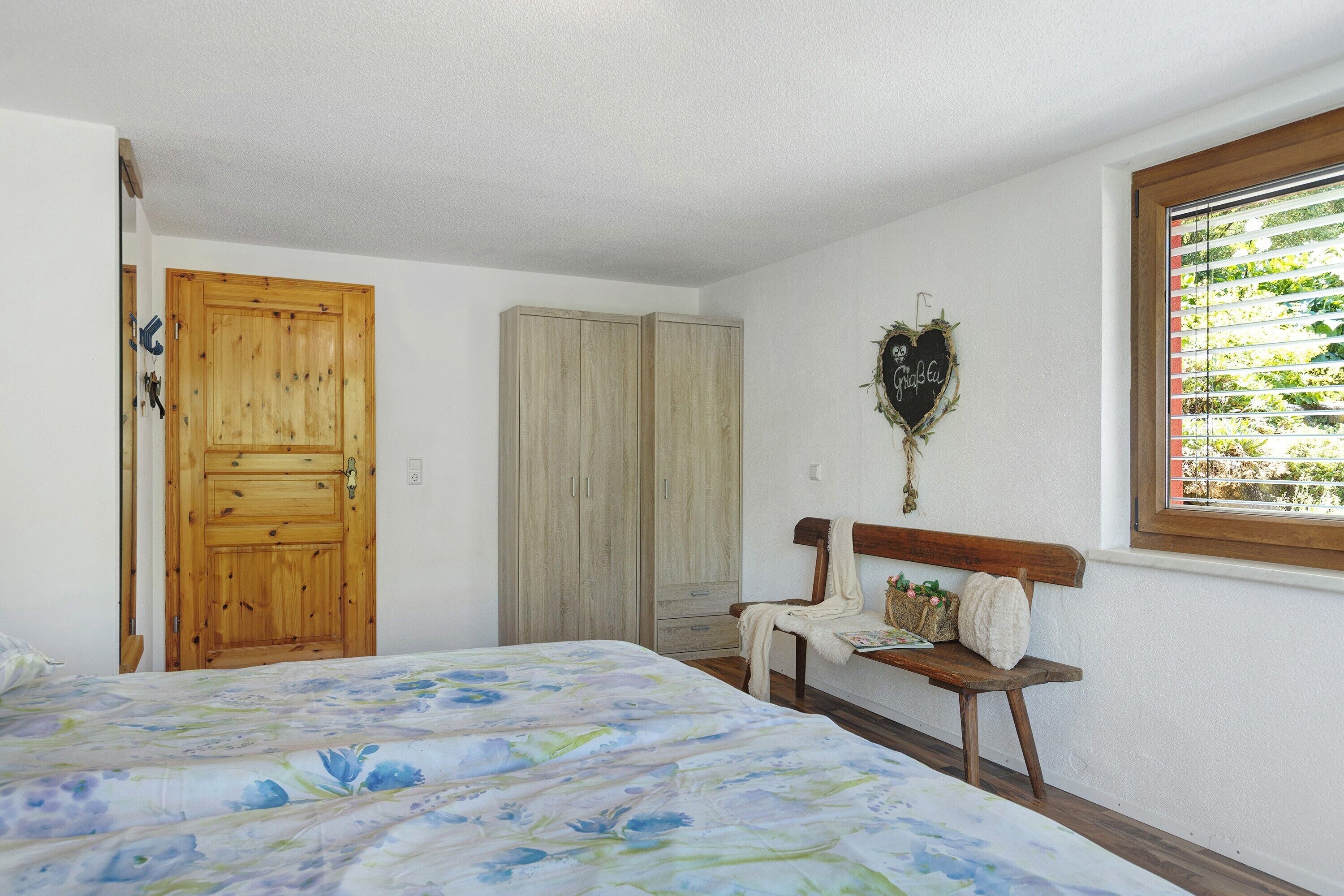 Een slaapkamer met een bed, een houten garderobe, een bankje en een raam met zonwering.