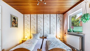 Una cama doble con cabecera de madera, dos mesitas de noche con lámparas, papel tapiz floral, una ventana con vista a vegetación y una planta colgante.