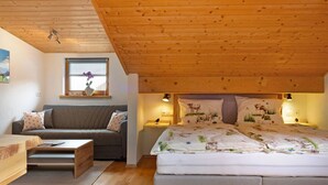 Een slaapkamer met een houten plafond, een bed met bloemenbeddengoed, een grijze bank, een klein tafeltje en een raam met uitzicht op de omgeving.