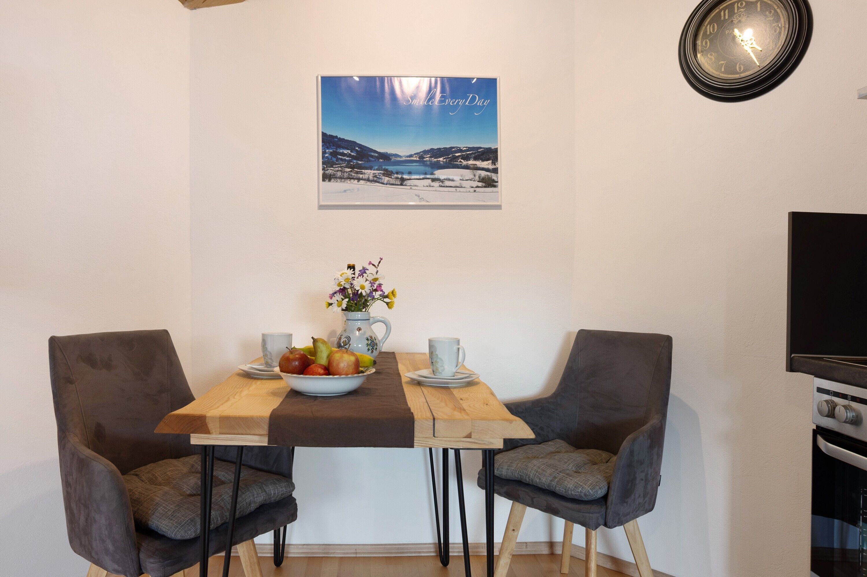 Un coin repas avec une table en bois, deux chaises grises à revêtement rembourré, un saladier de fruits, un vase avec des fleurs et un tableau encadré au mur.