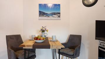 Un coin repas avec une table en bois, deux chaises grises à revêtement rembourré, un saladier de fruits, un vase avec des fleurs et un tableau encadré au mur.