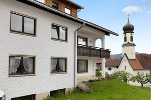Ein weißes Gebäude mit mehreren Fenstern, einem Balkon und einer Kirche mit einem Glockenturm im Hintergrund.
