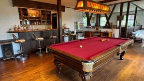 Une table de billard recouverte d’un drap de feutre rouge, un bar avec des tabourets et une grande fenêtre donnant sur l’extérieur.