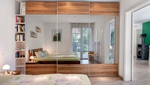 A bedroom with a wooden headboard, a bed with floral bedding, a bedside lamp, a bookshelf, and a view through a glass door.