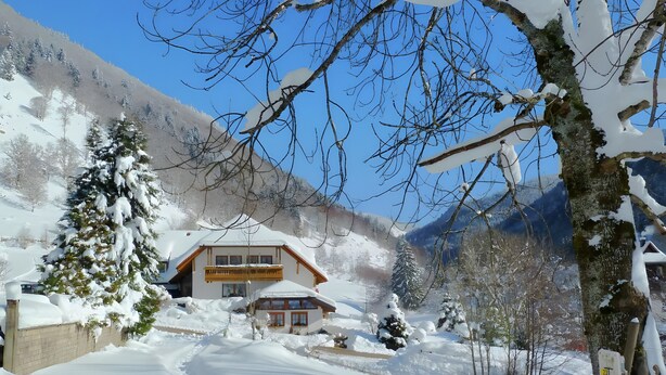 Una casa de montaña cubierta de nieve con fachada de madera, rodeada de árboles cargados de nieve y un cielo azul despejado.