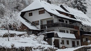 Un immeuble à plusieurs étages, dont le toit et les balcons sont recouverts de neige, entouré d’arbres chargés de neige et d’un mur de pierre.