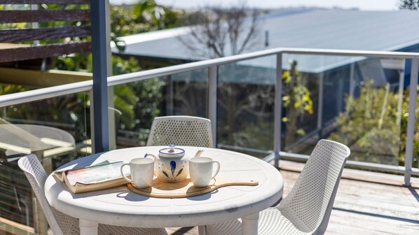 A round table with a white tablecloth, two white cups, a teapot, and a book on a balcony with a glass railing. - Angourie Blue 3 - Lifestyle Yamba (ANGOURIE)