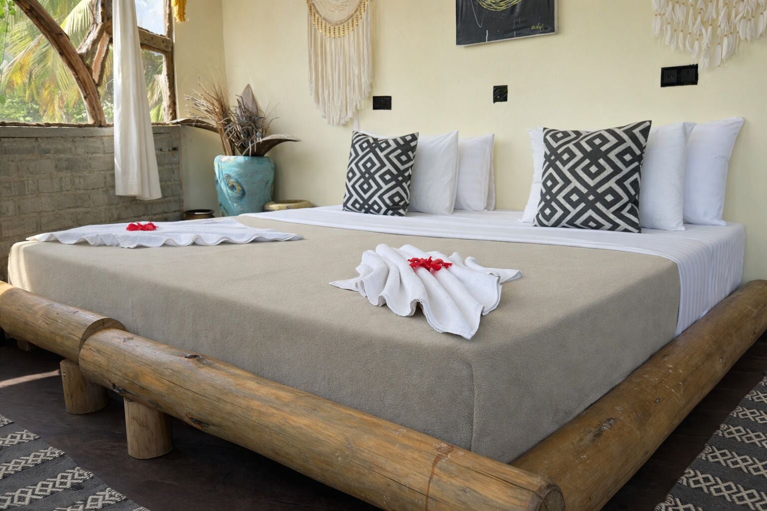Cama doble con almohadas blancas y estampadas, cabecero de madera y una planta decorativa en un jarrón azul.
