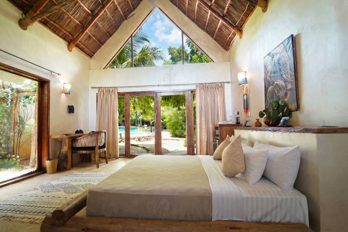 A spacious bedroom with a large bed, a desk, and a view of a pool and garden.