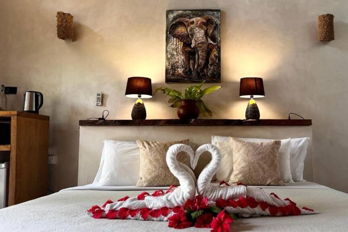 A neatly made bed with a heart-shaped towel arrangement, a painting of elephants on the wall, and two lamps on a shelf.