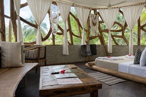 A room with a wooden table, a sofa, a TV, and a view of palm trees through a wooden-framed window. - The Nest Boutique Resort (Paje)