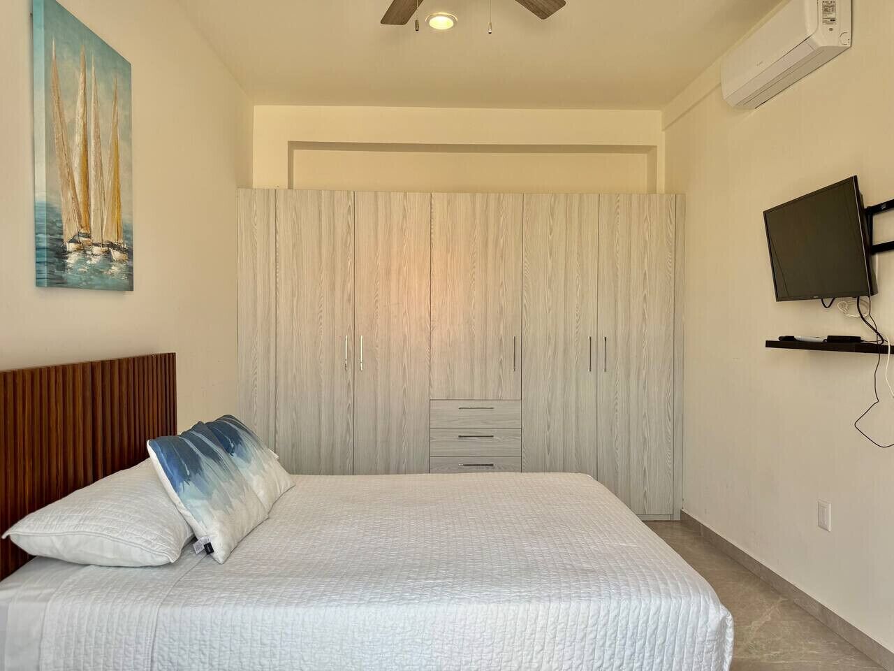 Een slaapkamer met een bed, een plafondventilator, een aan de muur bevestigde televisie en een schilderij van zeilboten.