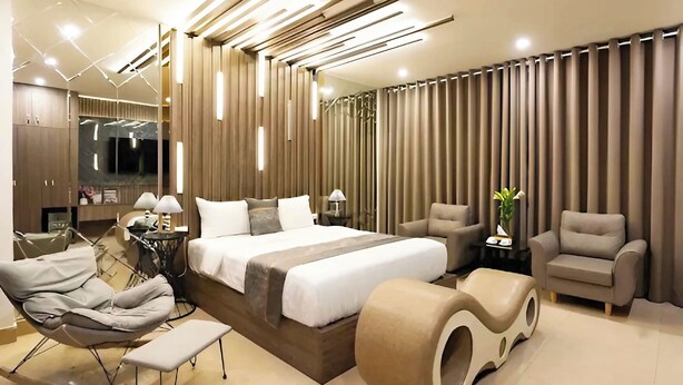 A modern hotel room with a large bed, a seating area with a chair and bench, a small table with a lamp, and a large mirror. - GRAD Queen Luxury Hotel (Hanoi)