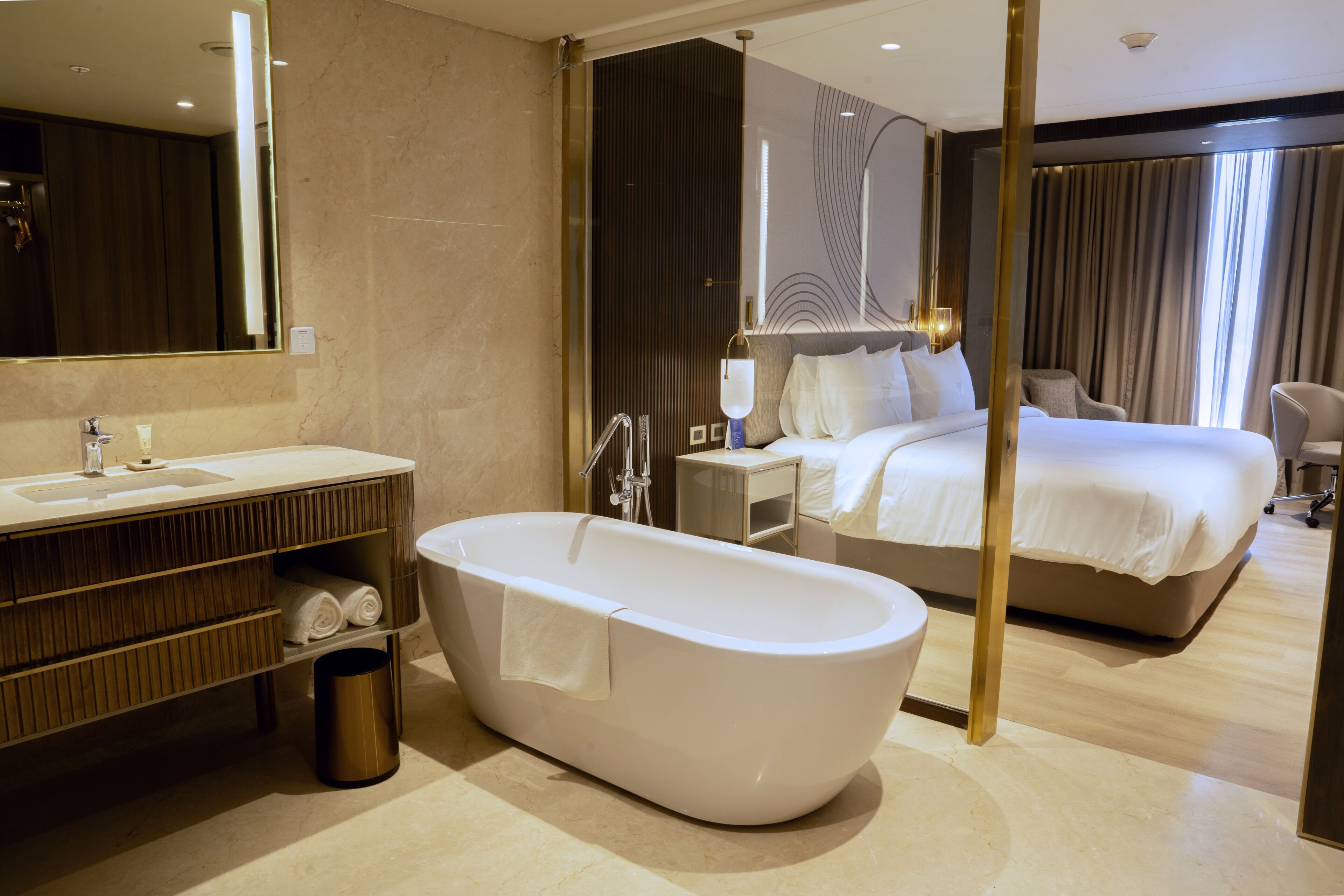 A modern hotel room with a large bathtub, a separate bathroom with a vanity, a bed with white linens, and a large mirror.