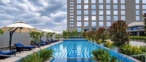 Una piscina con sillas de descanso y sombrillas, rodeada de follaje, con un edificio al fondo.