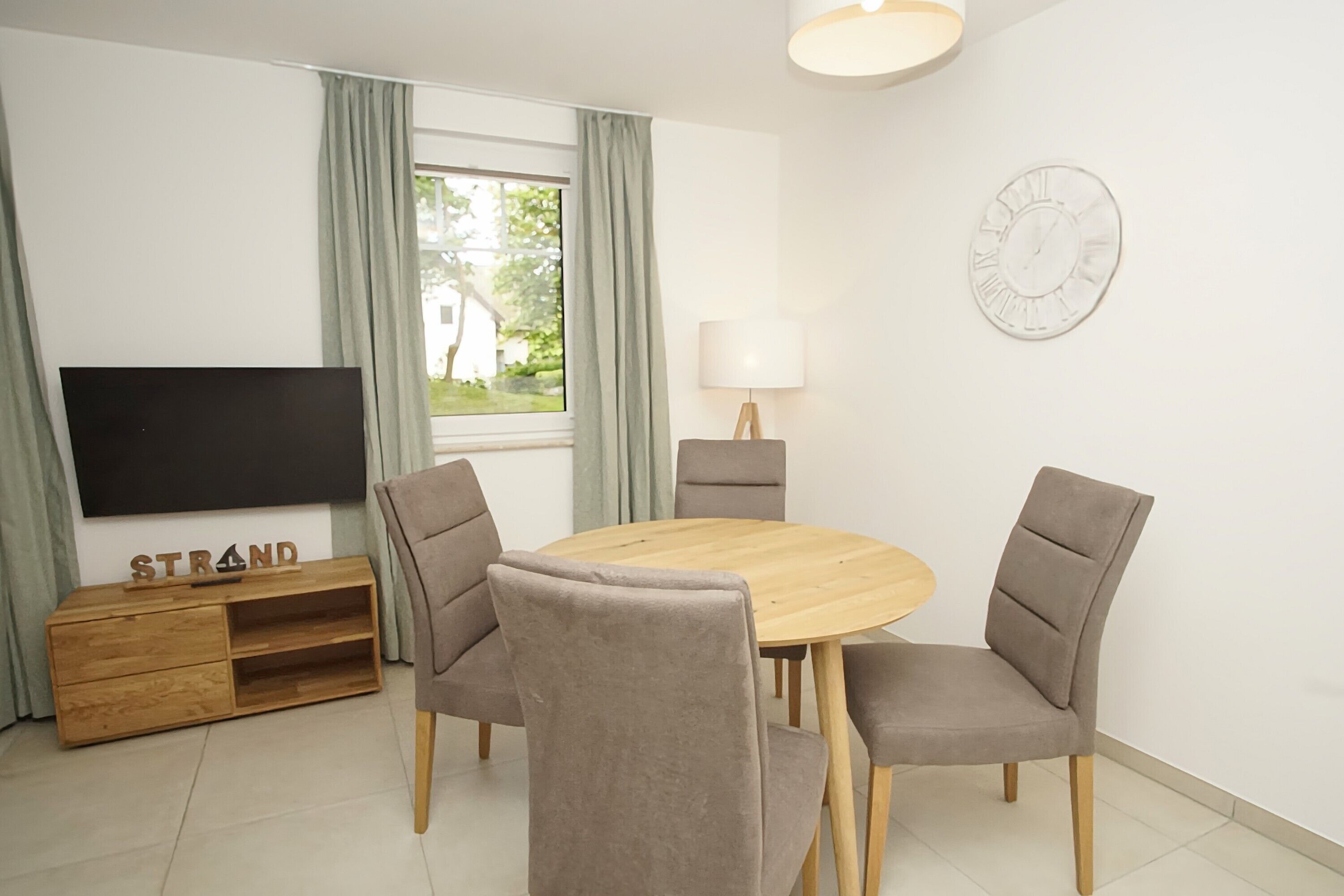 Un salon moderne avec une table à manger en bois, deux fauteuils gris à revêtement en tissu, un téléviseur à écran plat posé sur un meuble en bois, une lampe et une fenêtre avec des rideaux.