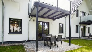 Une terrasse couverte avec une table et des chaises, un bâtiment blanc aux fenêtres à encadrement noir et une pelouse verdoyante.