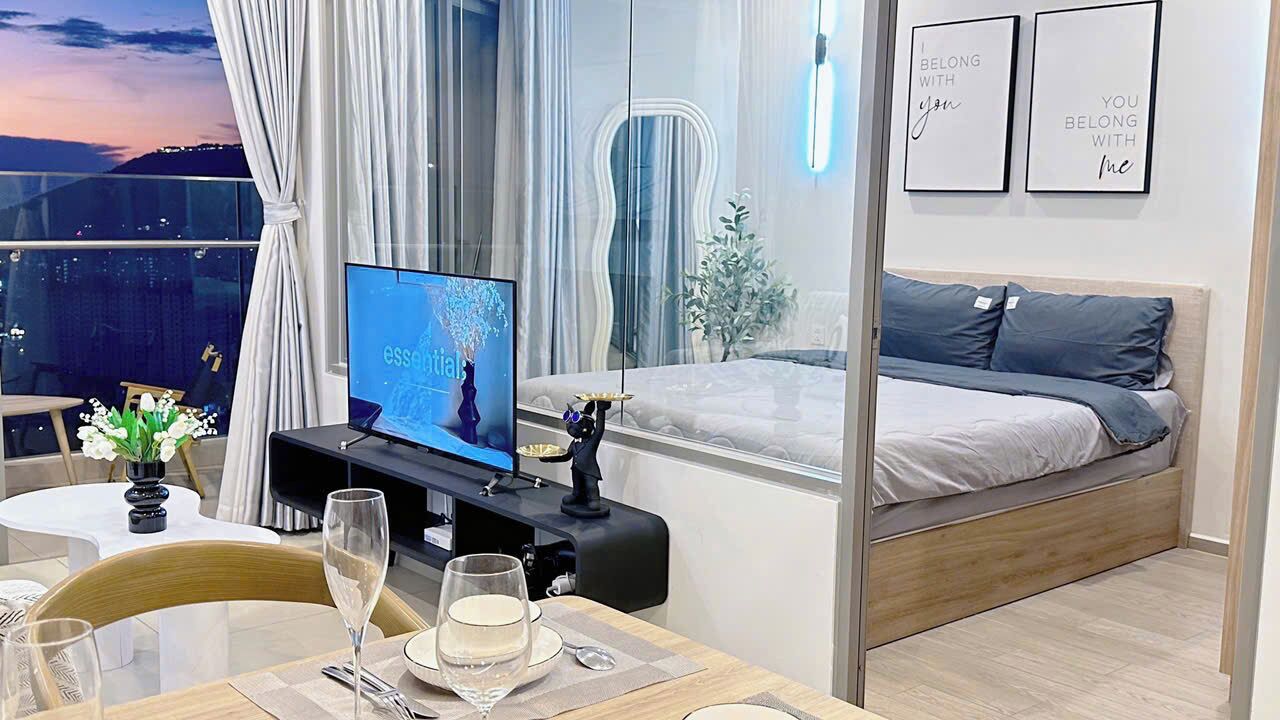 A modern hotel room with a balcony, a TV, a dining table with glasses, and a bed with pillows.