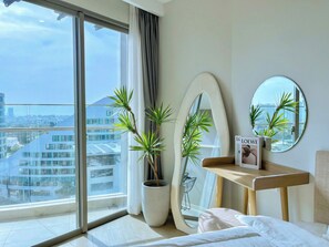 Una habitación de hotel moderna con un ventanal grande, una planta en maceta, un espejo redondo y una mesita de noche con un libro.