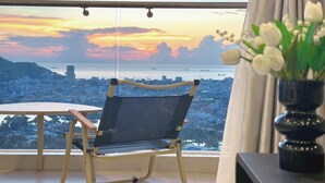 Habitación con vista a un paisaje urbano al atardecer, una silla, una mesita y un florero con flores.