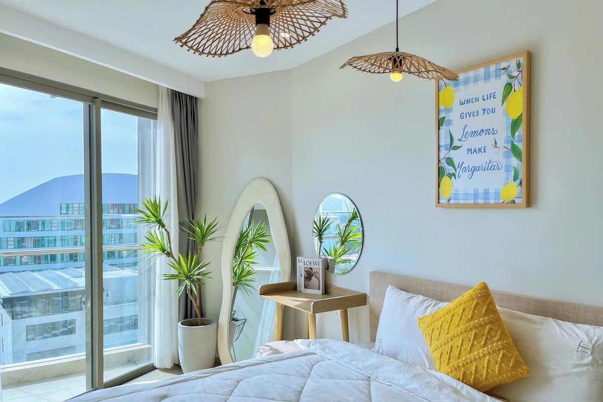 A modern hotel room with a large bed, two hanging wicker lamps, a large window with a view of buildings, a bedside table with a mirror, and a wall art piece.