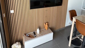 A modern living room with a flat-screen TV mounted on a wooden slatted wall, a small seating area with a vase and remote controls, and a dining area with a table and chairs. - Seemingly cosy, close to the beach and in the heart of the golf course (Saint-Cyprien)