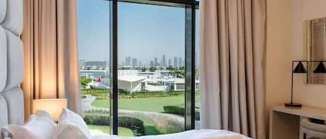 A hotel room with a large window, a bed with white linens, a desk with a lamp, and a view of a cityscape and golf course.