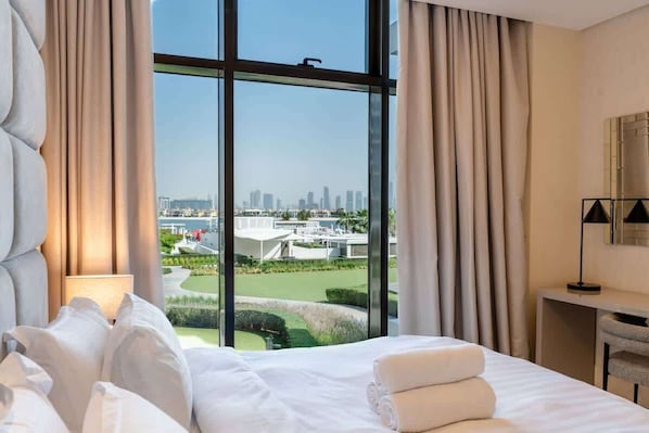 A hotel room with a large window, a bed with white linens, a desk with a lamp, and a view of a cityscape and golf course.