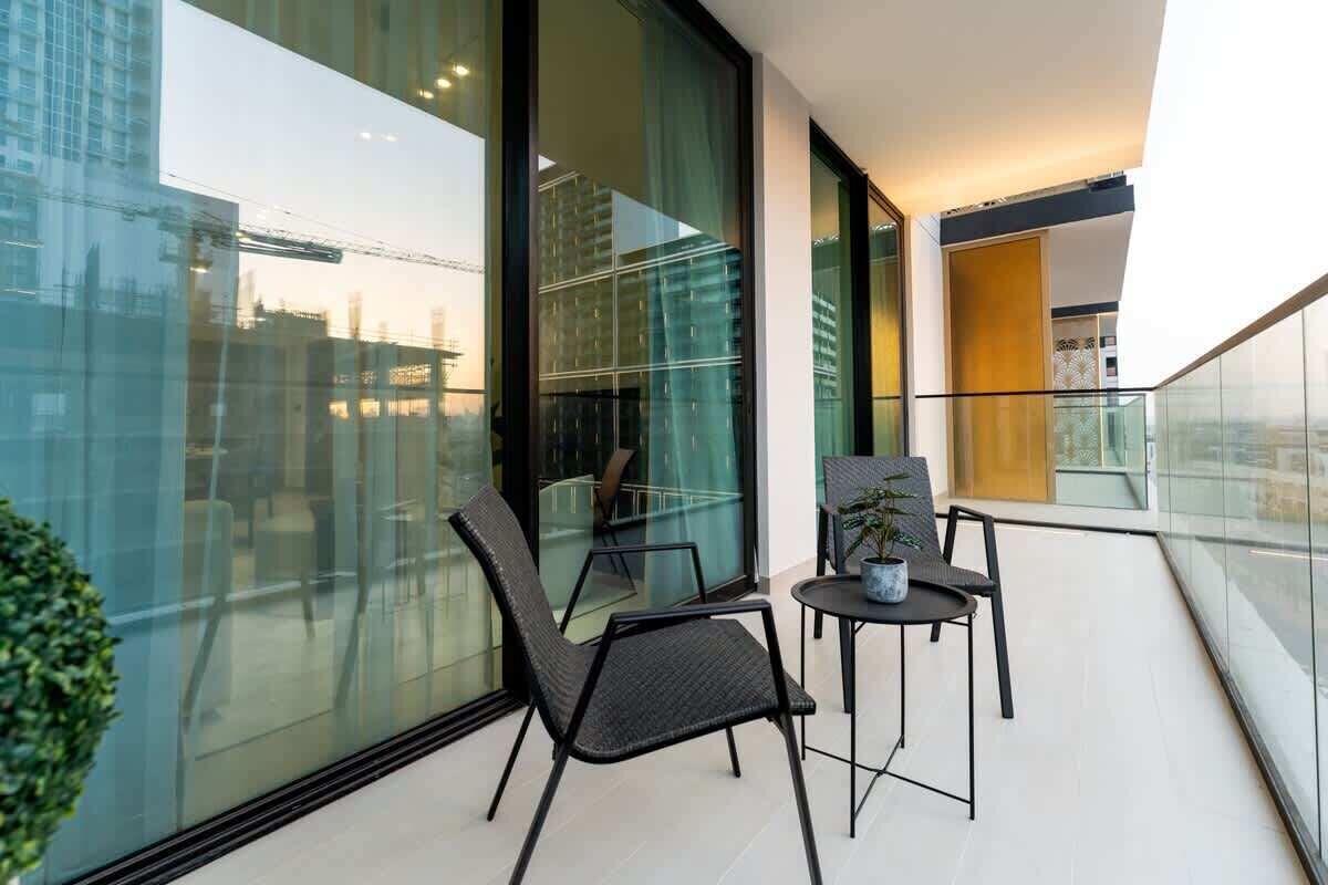 A balcony with two chairs and a small table, a potted plant, and a view of a cityscape through large glass windows.