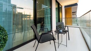 Un balcon avec deux chaises et une petite table, une plante en pot, et une vue sur le paysage urbain à travers de grandes fenêtres en verre.