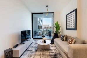 Una sala de estar moderna con un televisor de pantalla plana, una mesa de centro redonda, un sofá con cojines estampados y un amplio ventanal con vistas a edificios.