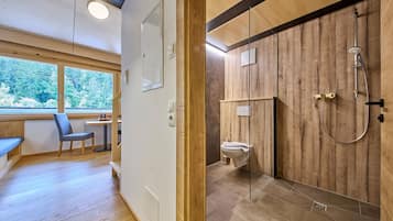 A modern bathroom with a wooden shower enclosure, a toilet, and a bench in a room with wooden flooring and a large window.