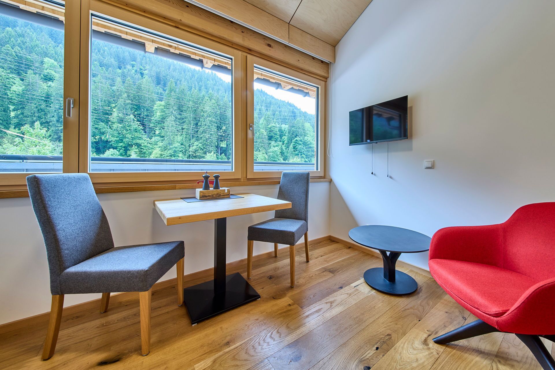 A room with a wooden table and chairs, a TV mounted on the wall, and large windows offering a forest view.