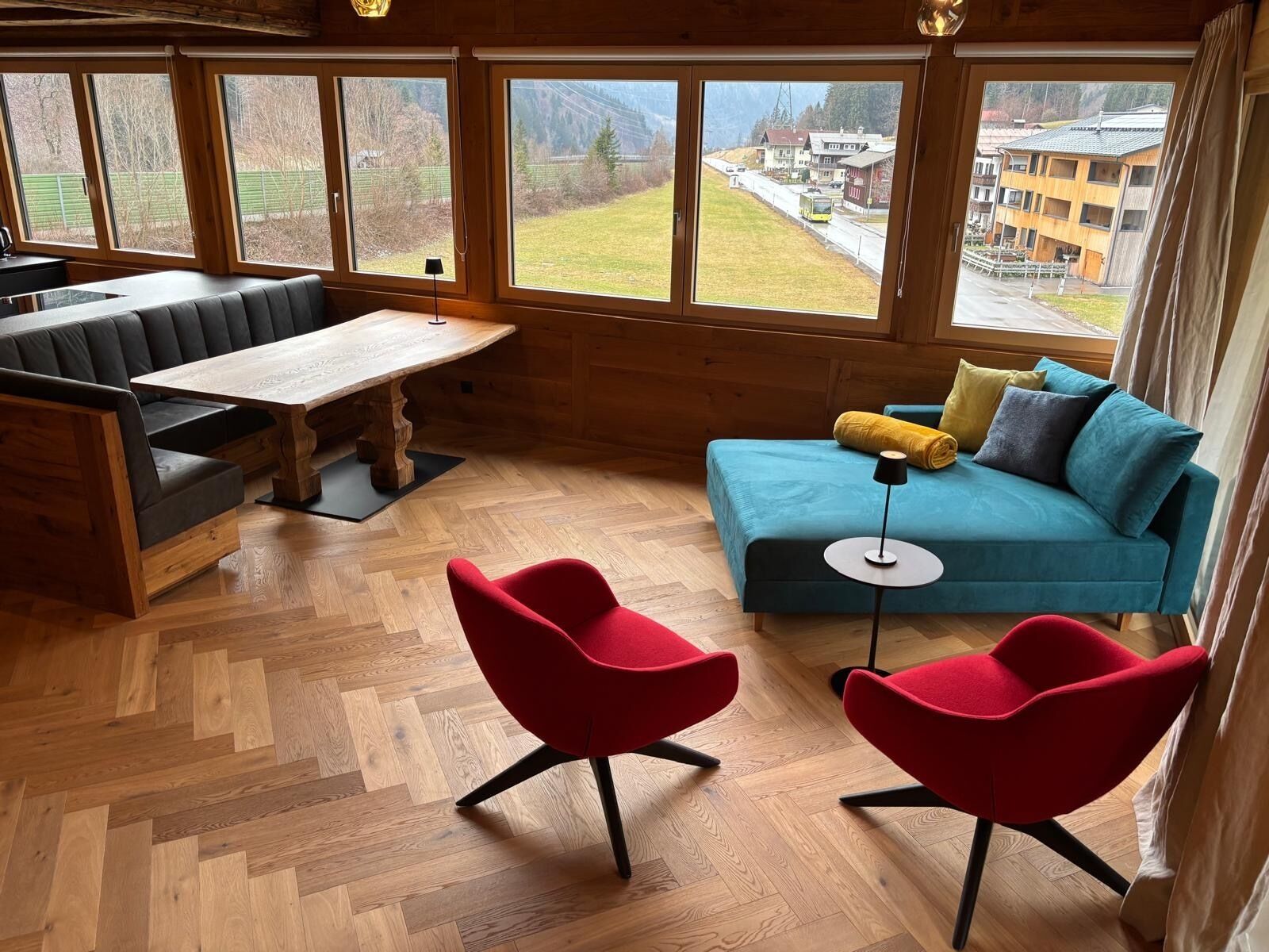 Una habitación acogedora con suelo de madera en espiga, un sofá verde azulado, una silla roja, un banco de madera y grandes ventanales con vistas a un pueblo y montañas.
