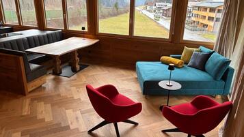 Una habitación acogedora con suelo de madera en espiga, un sofá verde azulado, una silla roja, un banco de madera y grandes ventanales con vistas a un pueblo y montañas.