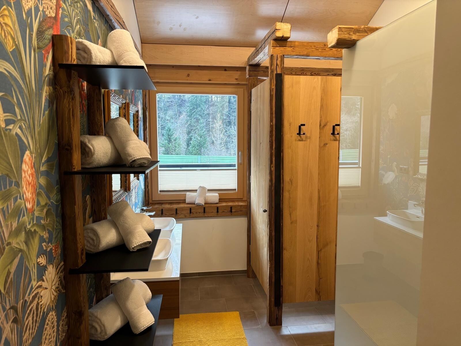 A bathroom with wooden shelves holding towels, a window with a view of trees, and a wooden door.