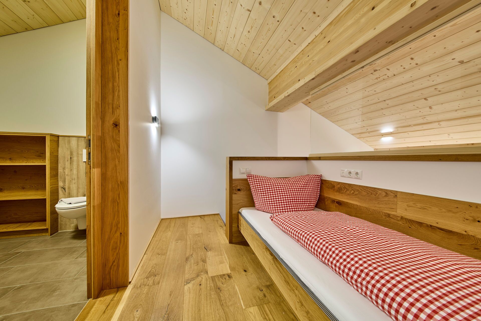 Een kleine kamer met een enkel bed, een houten hoofdbord en een geruit beddegoed. Links is een houten plankunit te zien, en rechts een witte muur met een lichtschakelaar.