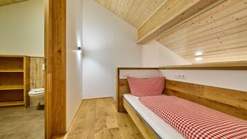 Een kleine kamer met een enkel bed, een houten hoofdbord en een geruit beddegoed. Links is een houten plankunit te zien, en rechts een witte muur met een lichtschakelaar.