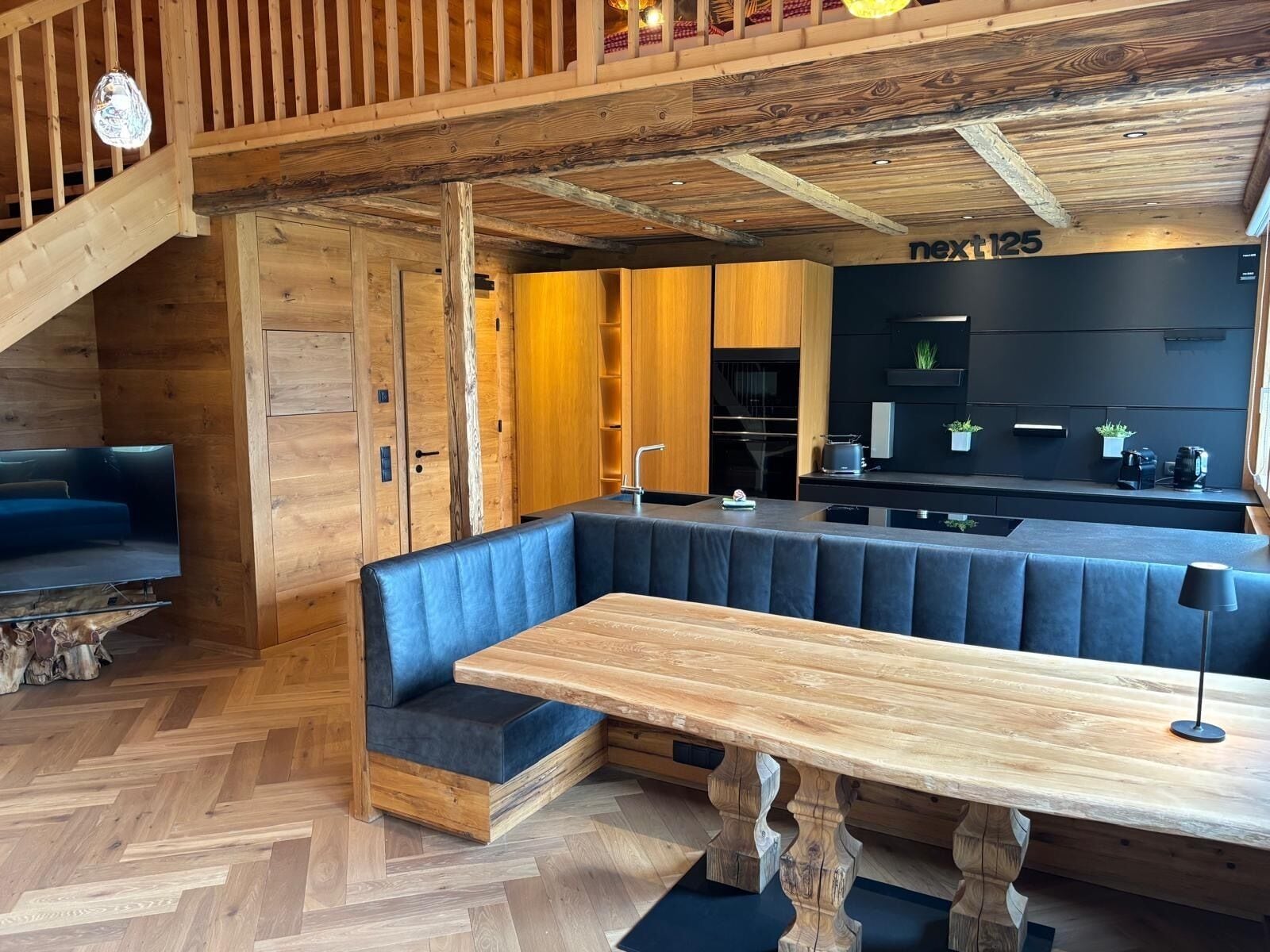 Een houten interieur met een grote houten tafel, blauw gestoffeerde zittingen en een keukenhoek.