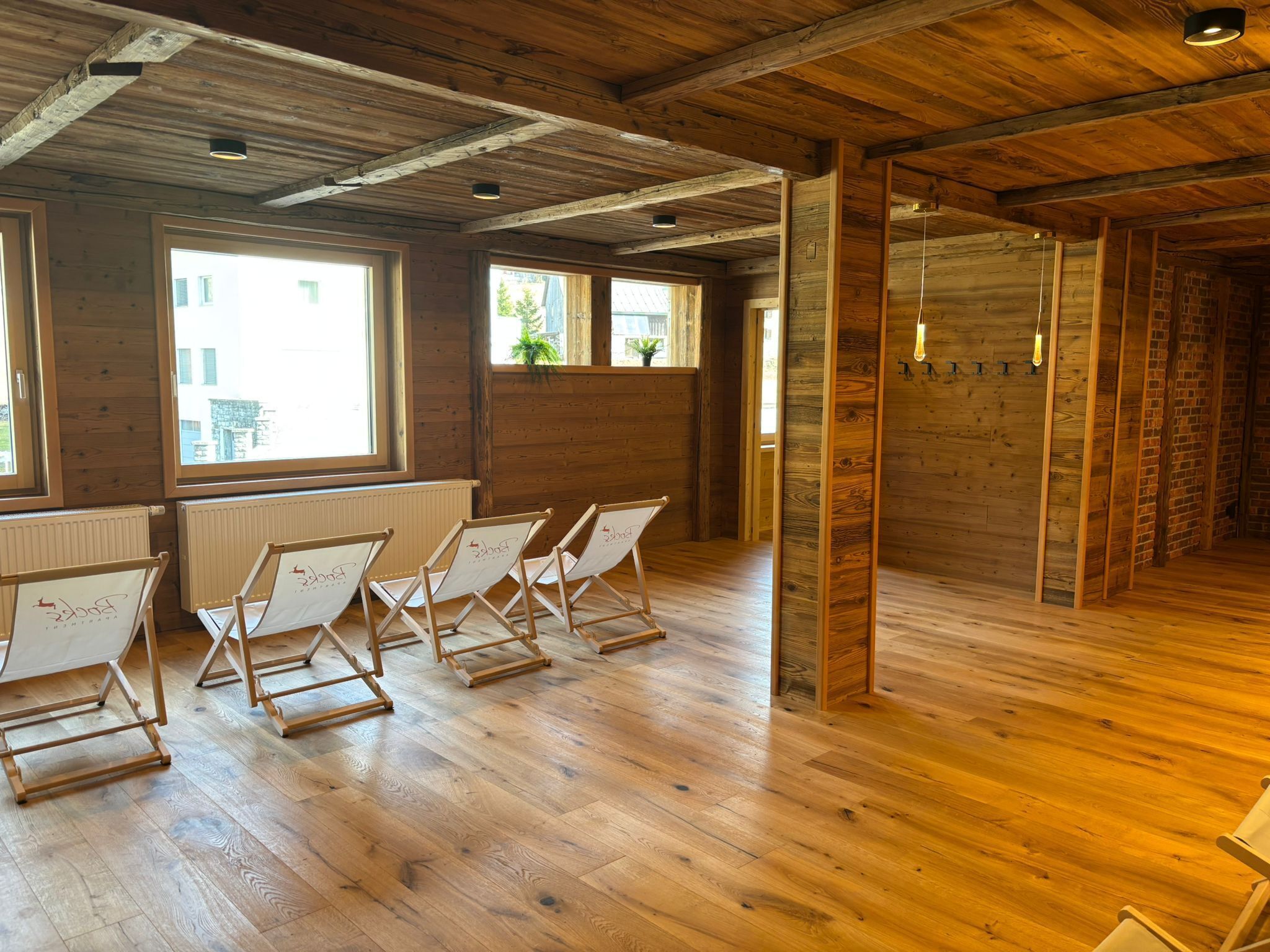 Une pièce avec un plancher en bois, de grandes fenêtres et un mur en briques. On y trouve quatre chaises pliantes, chacune portant un logo.