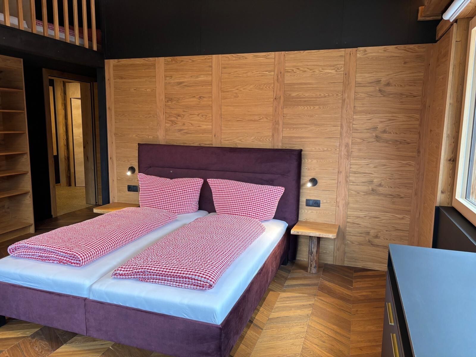 A bedroom with a wooden panel wall, a purple upholstered bed, checkered bedding, two bedside tables, and a herringbone-patterned floor.