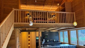 A modern loft-style interior with a wooden staircase, a mezzanine with a bed, a kitchen area, and a dining space with a bench. - Bocks Apartments 53C (Dalaas)