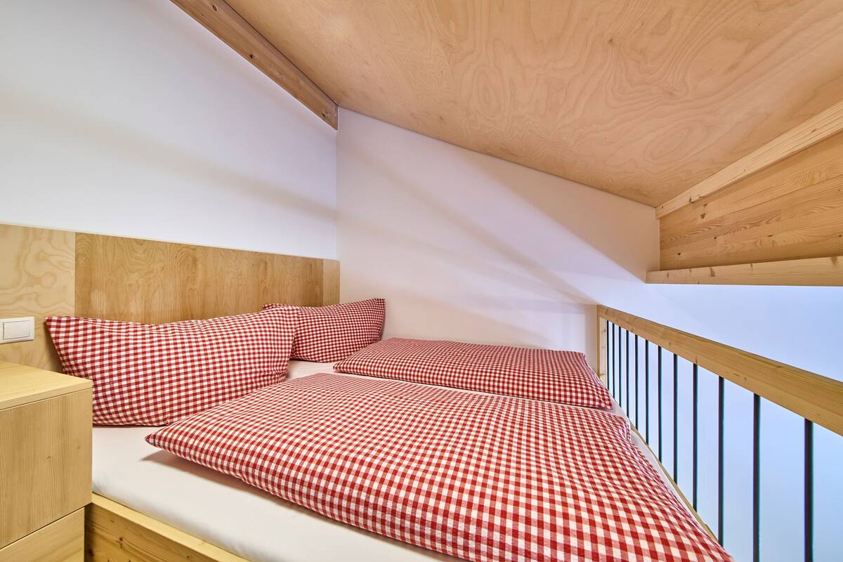 Une petite chambre à coucher accueillante, avec une tête de lit en bois, un lit simple recouvert d’une couette à carreaux, une table de chevet et une fenêtre donnant sur le ciel.
