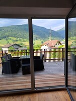 Terraza de madera con mobiliario de mimbre y vistas a un paisaje montañoso.