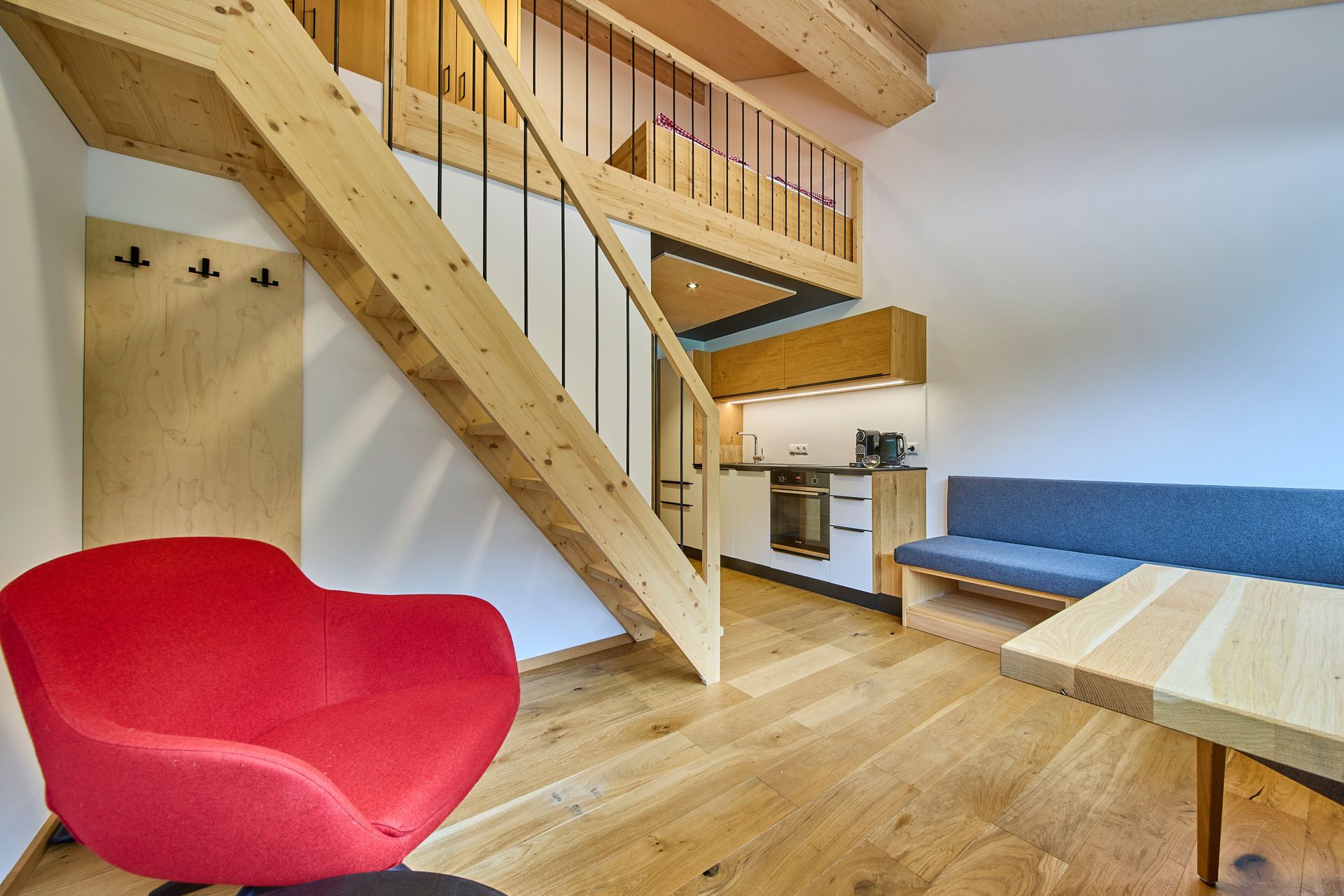 Une pièce de style loft avec un escalier en bois, un coin cuisine, une chaise rouge et un canapé bleu.