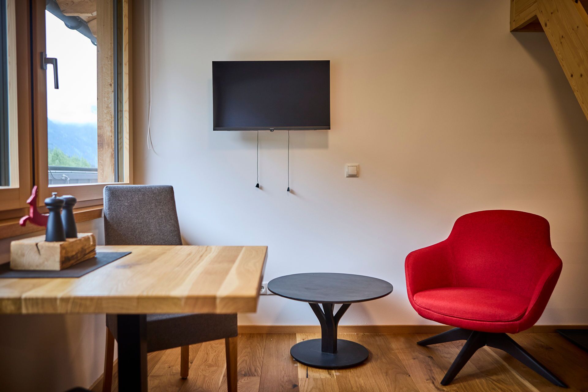 Une pièce avec une table en bois, une chaise rouge, une petite table ronde et un téléviseur fixé au mur.