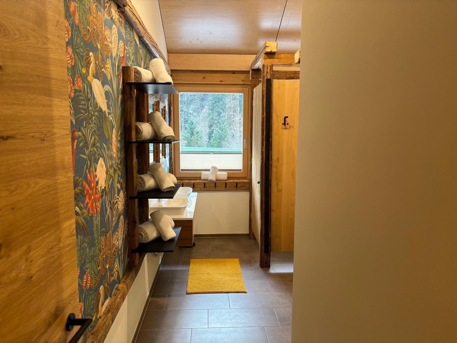 A narrow hallway with wooden walls, a window with a view of trees, a yellow rug, and shelves with towels.