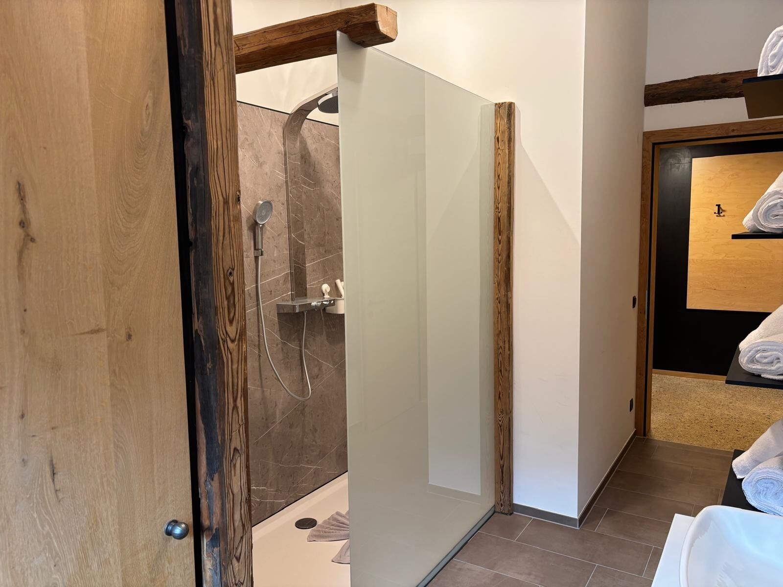 A modern bathroom with a glass shower enclosure, wooden accents, and a neatly made bed with towels.